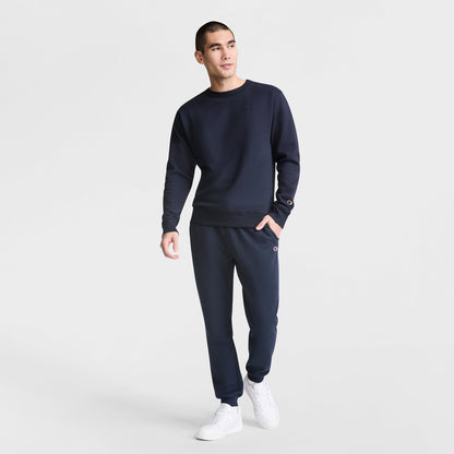 A man stands against a white background wearing the Champion Powerblend Crewneck Sweatshirt with matching sweatpants and white sneakers, looking slightly to the side in a relaxed pose.