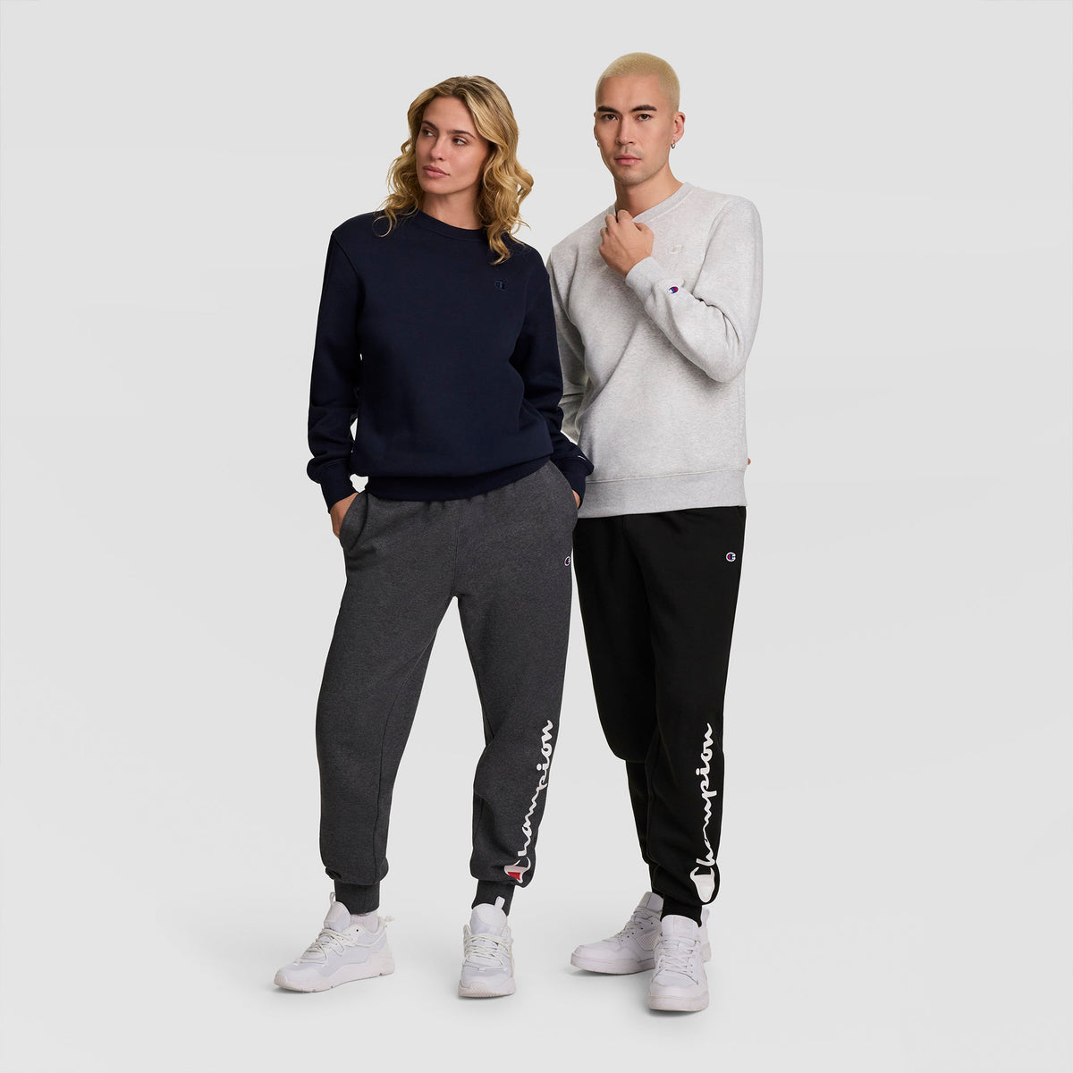 Two people stand side by side wearing casual Champion outfits. The person on the left sports a Champion Powerblend Crewneck Sweatshirt, C Logo in navy and dark gray, while the person on the right opts for a light gray and black Champion men’s sweatshirt crafted from soft athletic fleece. Both wear white sneakers.