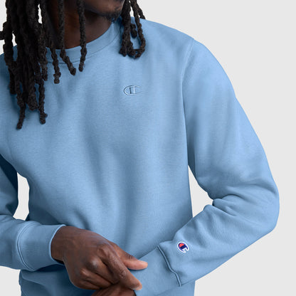 A person with long hair is wearing the Champion Powerblend Crewneck Sweatshirt, C Logo in light blue, featuring the classic athletic fleece design and embroidered Champion logos on the chest and sleeve, set against a plain light background.