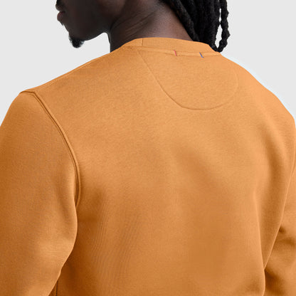 A close-up of a person wearing a mustard yellow Champion Powerblend Crewneck Sweatshirt, C Logo, shown from the back. The fabric texture and neckline details are visible, and the person has dark braided hair.
