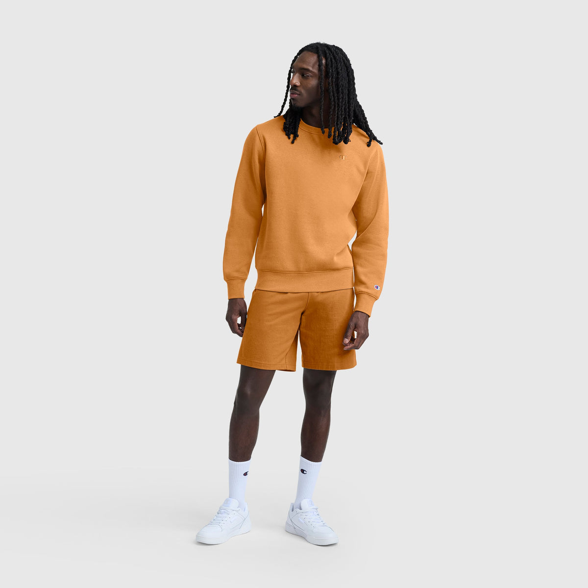 A man with long hair is wearing a matching orange Champion Powerblend Crewneck Sweatshirt with C Logo and shorts set, paired with white crew socks and white sneakers, standing against a plain light gray background.