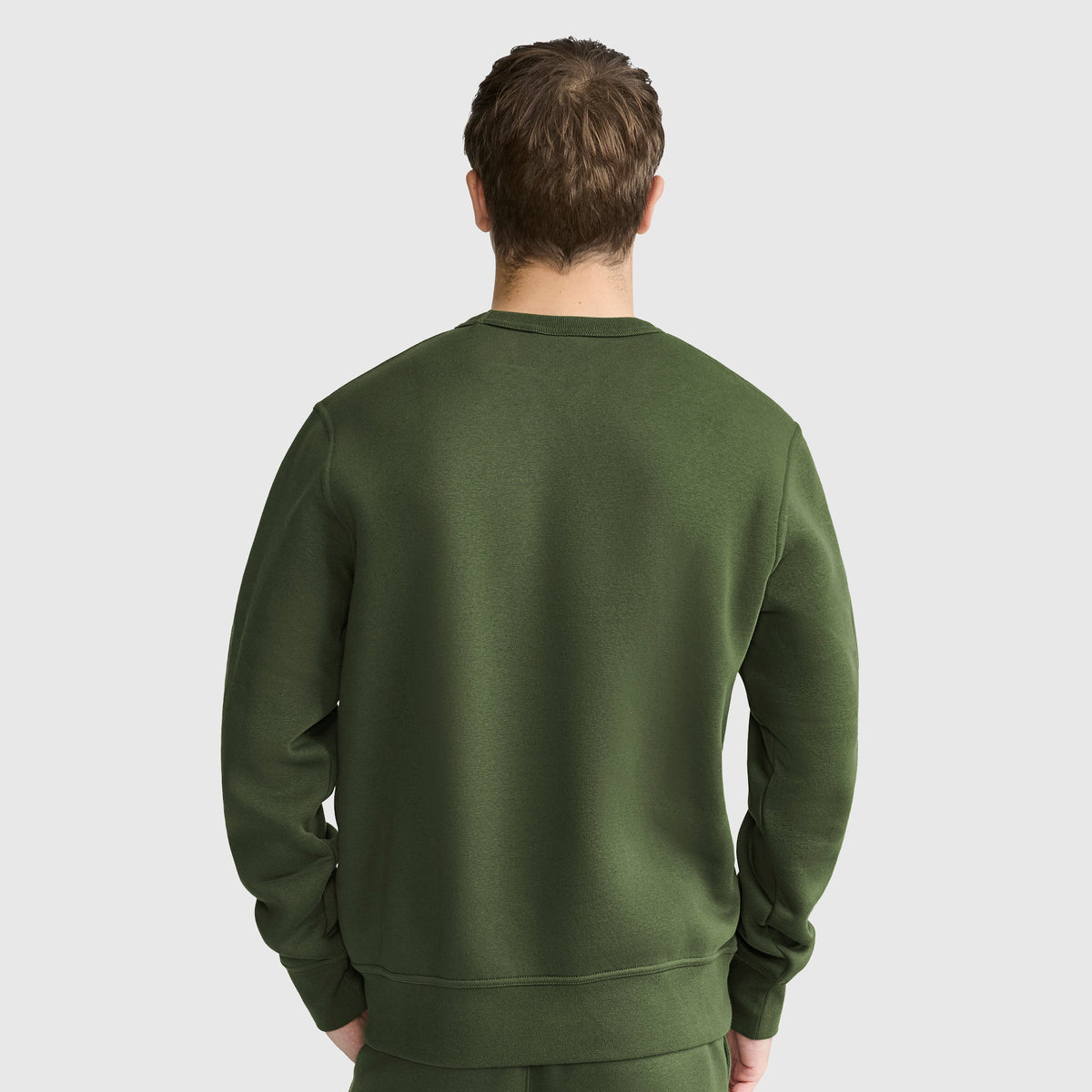 A person with short brown hair is shown from the back, wearing a plain dark green Champion Powerblend Crewneck Sweatshirt, C Logo, along with matching pants, standing against a light gray background.