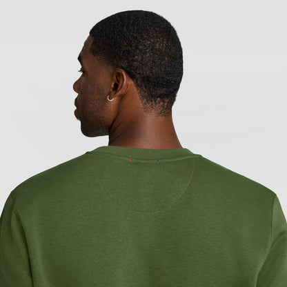 A man seen from behind wearing an olive green Champion Powerblend Crewneck Sweatshirt, C Logo - Big & Tall, with small red and blue stitch detailing at the back of the neck.