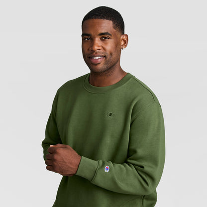 A man wears an olive green Champion Powerblend Crewneck Sweatshirt, C Logo - Big & Tall featuring a small chest logo and a sleeve cuff patch.