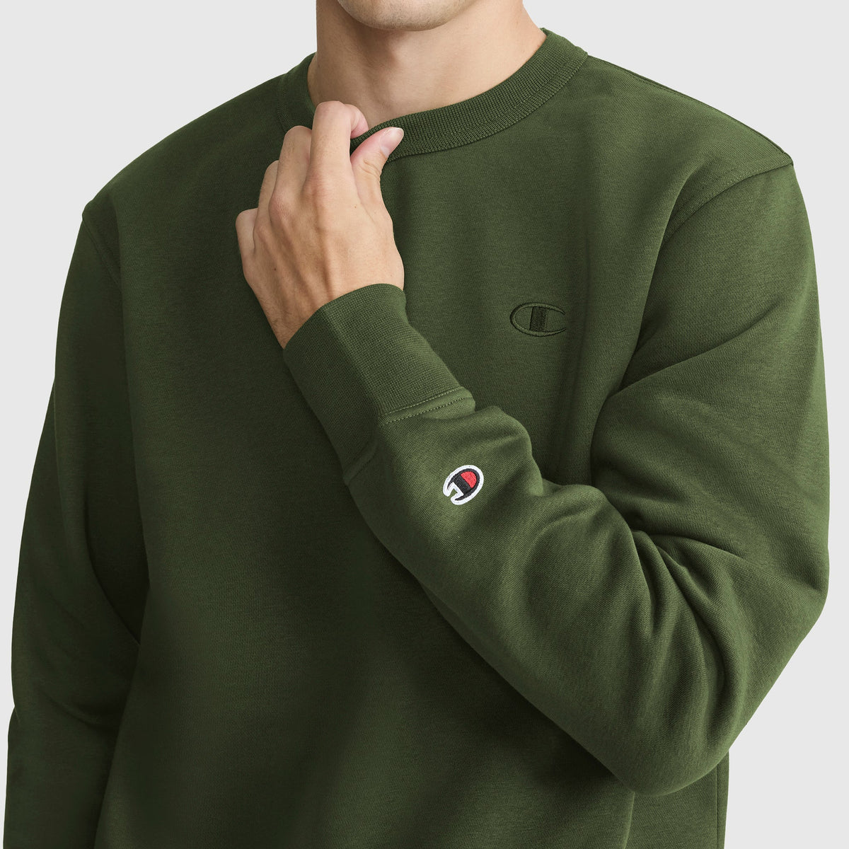 A person wearing a dark green Champion Powerblend Crewneck Sweatshirt, featuring the signature C logo embroidered on the chest and sleeve, stands against a plain light background.