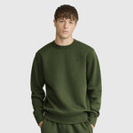 A young man with short brown hair and a neutral expression stands against a plain light gray background, wearing an olive green Champion Powerblend Crewneck Sweatshirt with a C logo and matching pants.