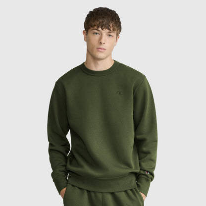 A young man with short brown hair and a neutral expression stands against a plain light gray background, wearing an olive green Champion Powerblend Crewneck Sweatshirt with a C logo and matching pants.
