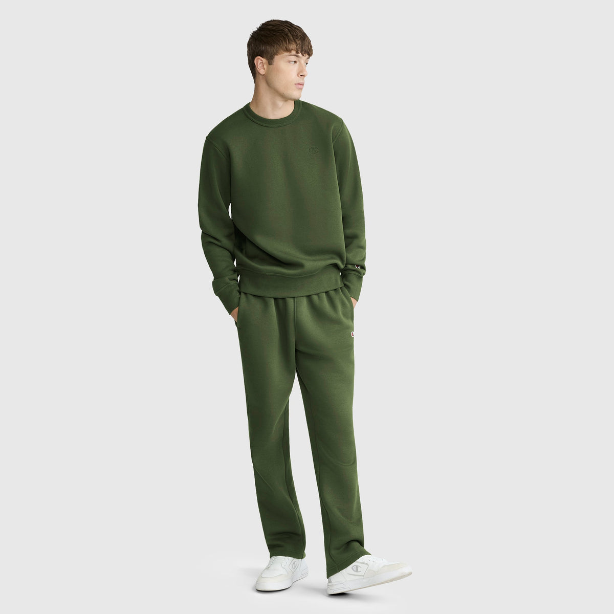 A young man is wearing a Champion Powerblend Crewneck Sweatshirt, C Logo, in olive green along with coordinating sweatpants. He stands with his hands in his pockets, looking to the side, and completes the look with white sneakers against a plain light gray background.