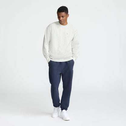 A man stands with his hands in his pockets, looking down in a minimalist white studio, wearing a Champion Reverse Weave Crewneck Sweatshirt with the C Logo, dark blue jogger pants, and white sneakers.