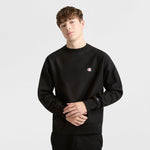 A young man with short brown hair wears a black Champion Reverse Weave Crewneck Sweatshirt, C Logo, made from heavyweight fleece. He stands against a plain light background, looking at the camera with a neutral expression.
