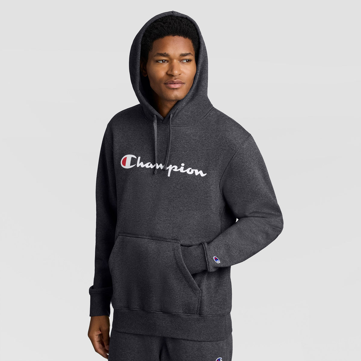 A man wearing a dark gray Champion Powerblend Hoodie, Full Script Logo with the brand name printed in white and red across the chest.