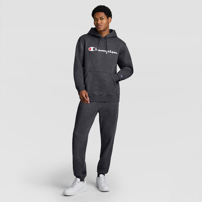 A person wearing a dark gray Champion Powerblend Hoodie, Full Script Logo, with matching sweatpants stands against a plain light gray background, white sneakers completing the outfit.
