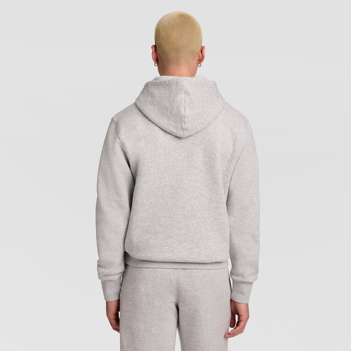 A person with short blond hair, wearing a light gray Champion Powerblend Hoodie featuring the full script logo and matching sweatpants, is standing with their back to the camera against a plain light background.