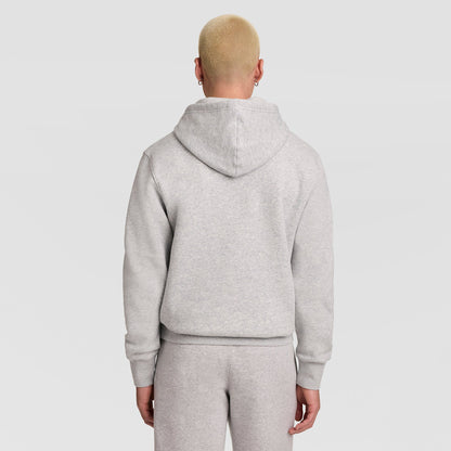 A person with short blond hair, wearing a light gray Champion Powerblend Hoodie featuring the full script logo and matching sweatpants, is standing with their back to the camera against a plain light background.