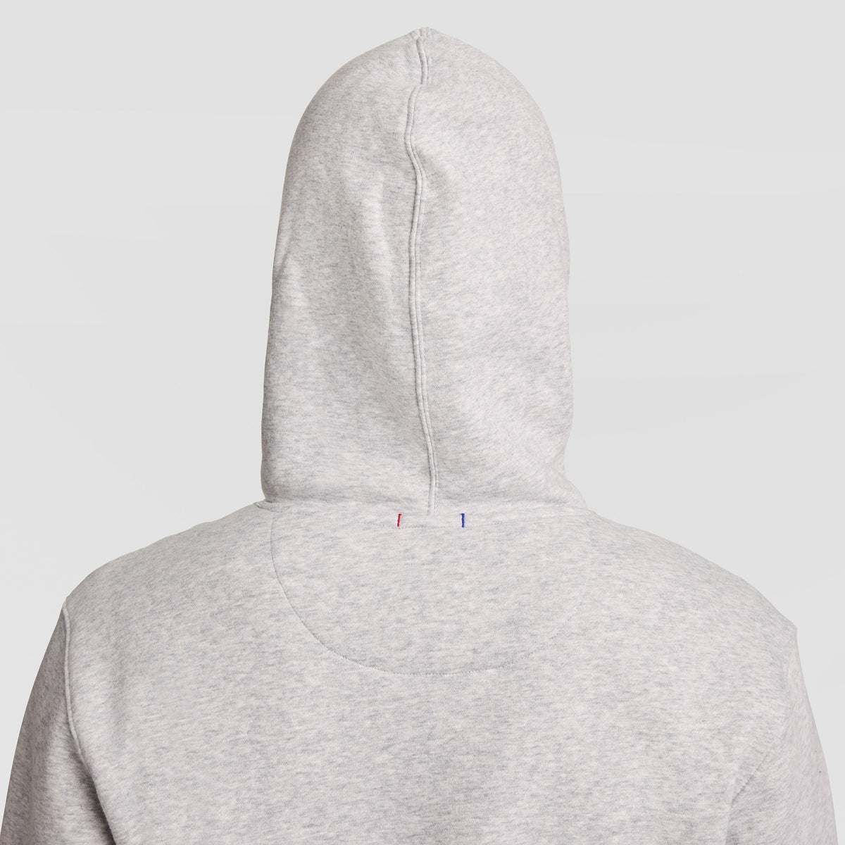 A person wearing a light gray Champion Powerblend Hoodie, Full Script Logo, with the hood up, shown from the back. Two small vertical stitch marks—one red and one blue—are visible on the upper part of the hood.