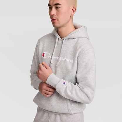 A person wearing a Champion Powerblend Hoodie, Full Script Logo in light gray with matching sweatpants stands against a plain white background, looking to the side with one hand near their chest.