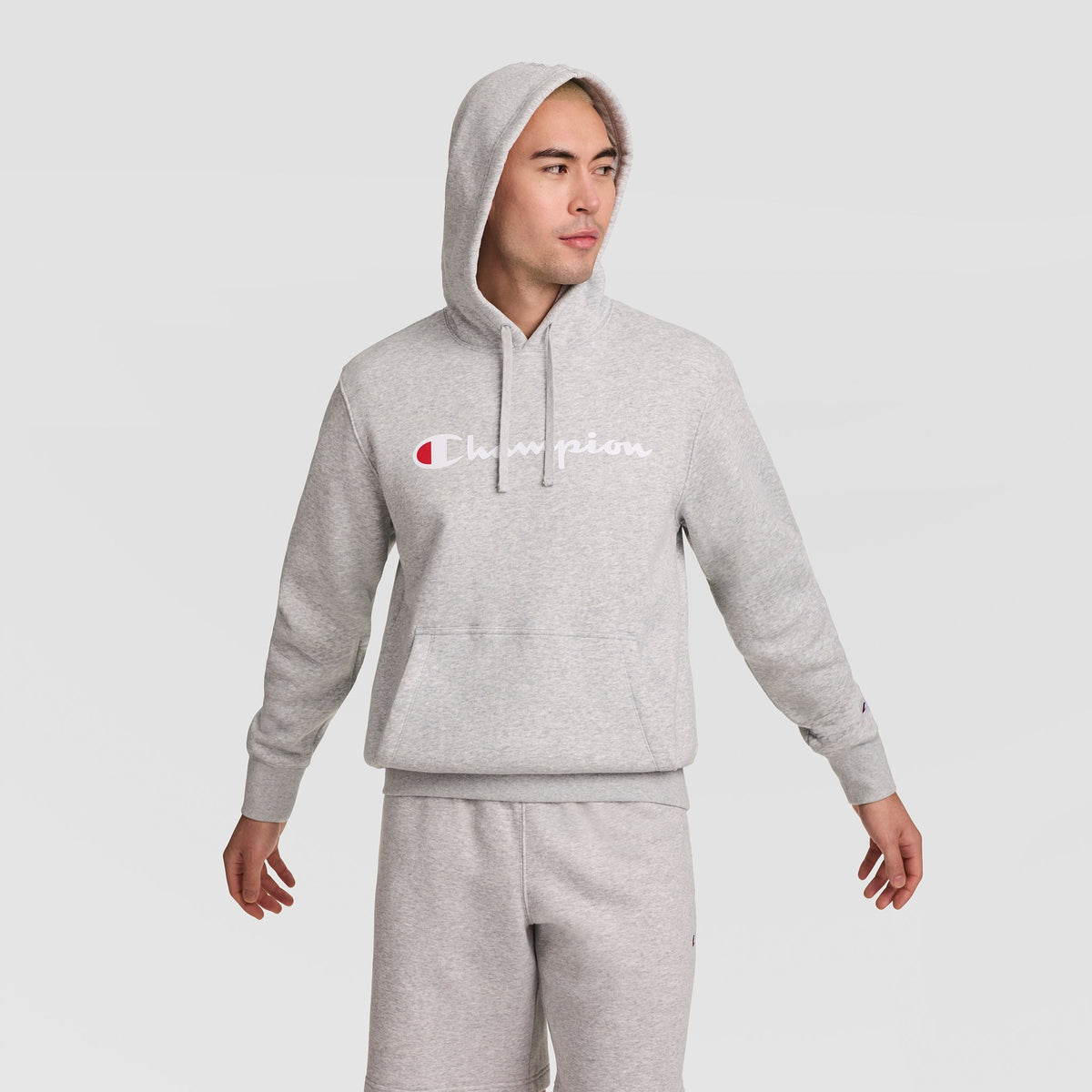A person wearing a light gray Champion Powerblend Hoodie with the Full Script Logo and matching sweatpants stands with arms slightly out, looking to the side against a plain light background.