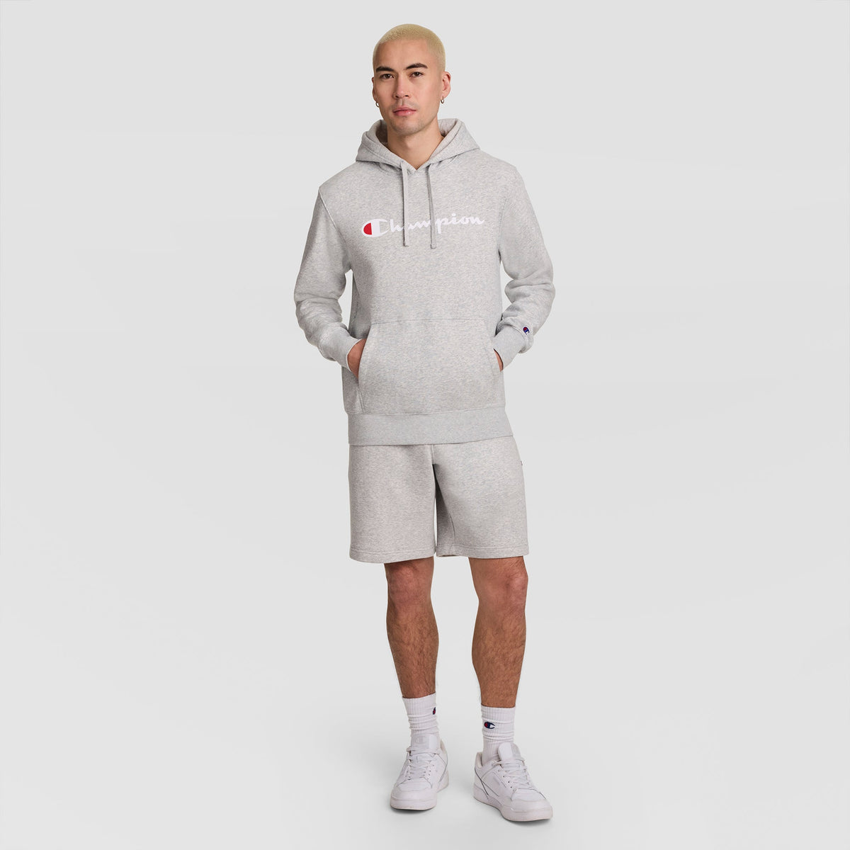 A person with blonde hair wears a gray Champion Powerblend Hoodie featuring the full script logo, along with matching shorts, white socks, and white sneakers, standing against a plain light gray background.