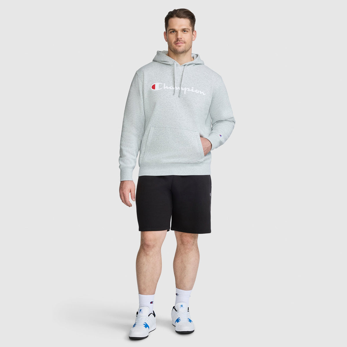 A man wearing a grey Champion Powerblend Hoodie, Full Script Logo - Big & Tall, black shorts, and white sneakers.