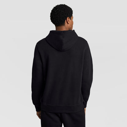 A person with short curly hair is standing with their back to the camera, wearing a Champion Powerblend Hoodie, Full Script Logo, paired with black pants against a light gray background.