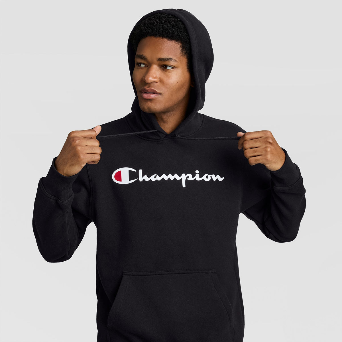 A man wearing a black Champion Powerblend Hoodie, Full Script Logo, featuring a red and white script logo across the chest.