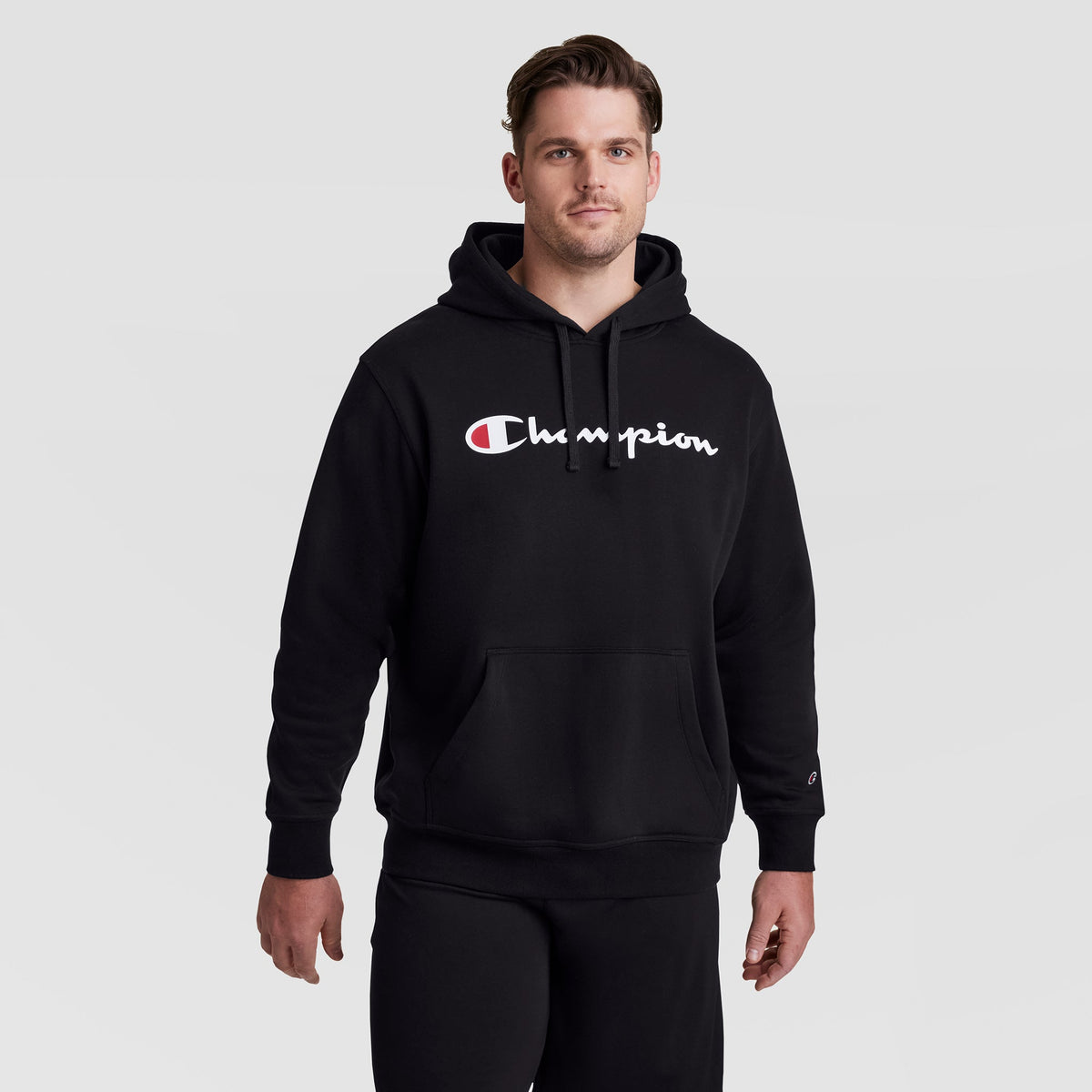 A man wearing a Champion Powerblend Hoodie, Full Script Logo - Big & Tall featuring a soft fleece pullover design with a large white and red logo on the chest.