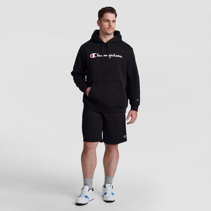A man wears a black Champion Powerblend Hoodie, Full Script Logo - Big & Tall, paired with matching black shorts, grey socks, and white sneakers.