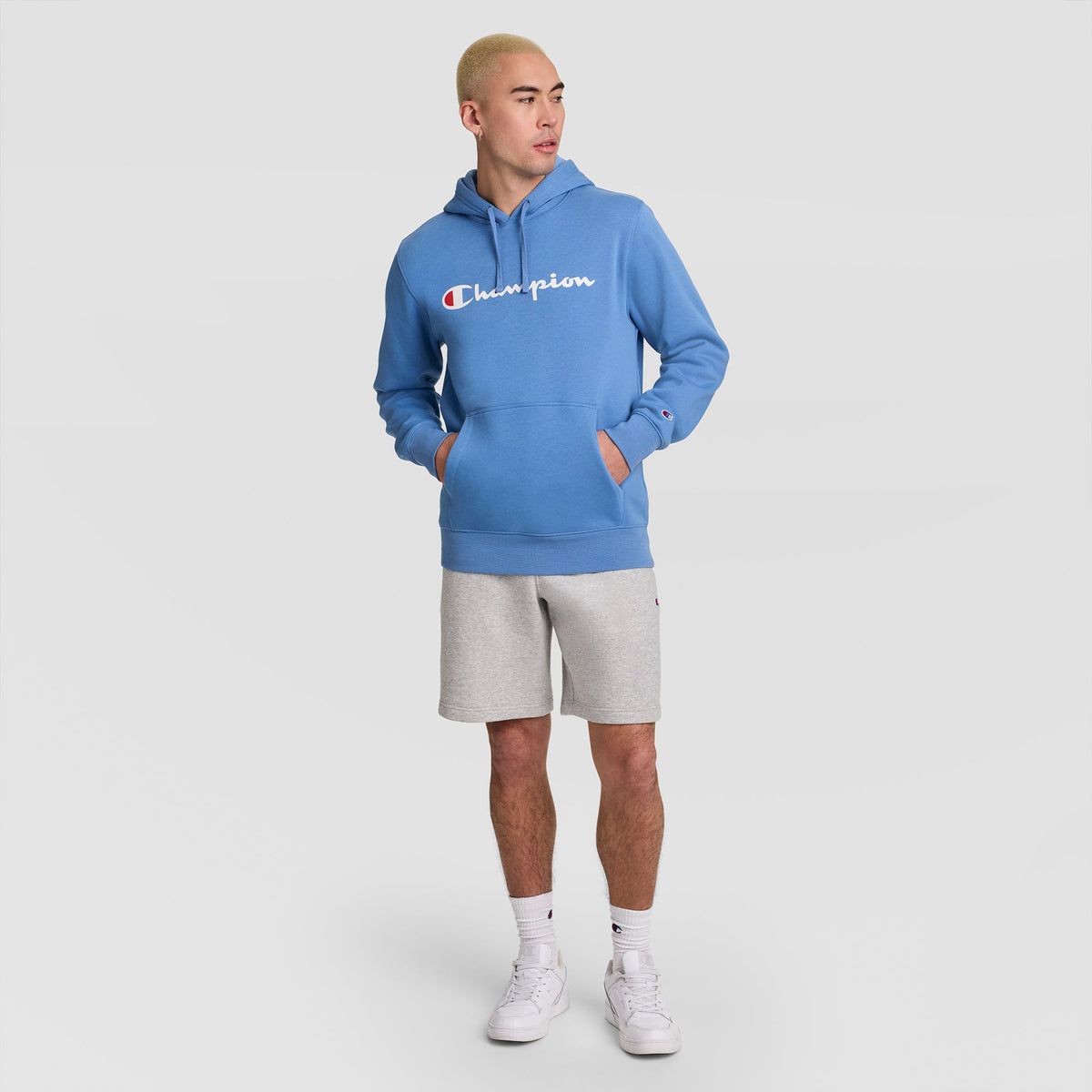 A person wearing a Champion Powerblend Hoodie, Full Script Logo in blue, paired with gray shorts, white socks, and white sneakers stands against a plain light background with hands in the hoodie pockets.