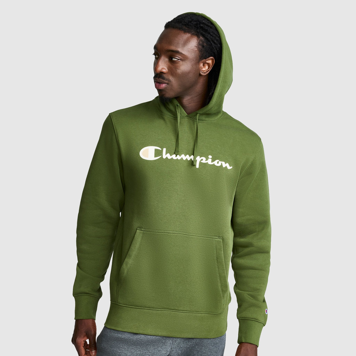 A man wearing a green Champion Powerblend Hoodie, Full Script Logo, featuring a front pocket and hood, looks to his left against a plain light gray background.