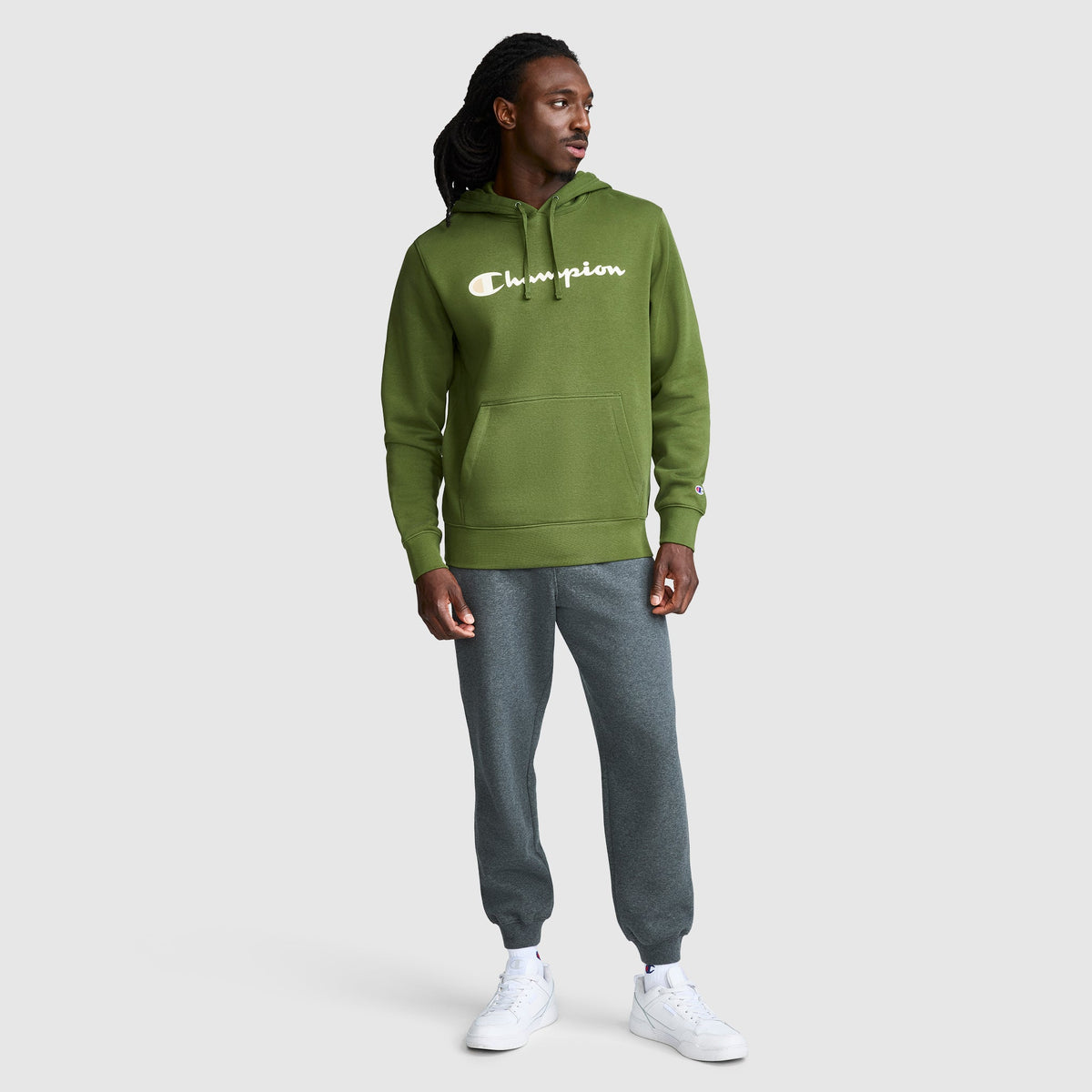A man wearing a Champion Powerblend Hoodie with the full script logo in green, paired with gray jogger pants and white sneakers, stands against a plain light gray background, looking to his left with one hand in his pocket.