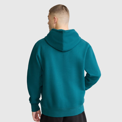 A person with short hair, wearing a teal Champion Powerblend Hoodie with a tonal script logo and black pants, stands facing away against a plain light gray background.