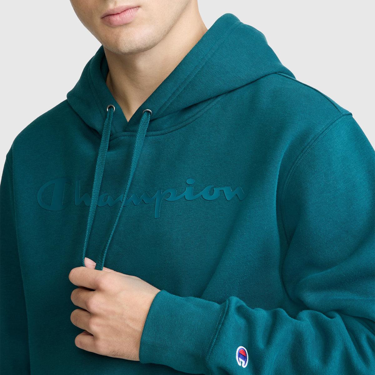 A person wearing a teal Champion Powerblend Hoodie with a tonal script logo embossed on the chest and a small Champion patch on the sleeve, holding the hoodie strings. Crafted from soft Powerblend Fleece by Champion for comfort and durability.