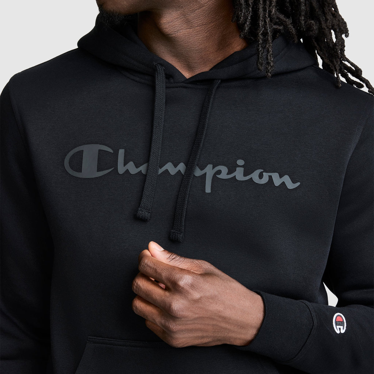 A person wearing a black Champion Powerblend Hoodie, Tonal Script Logo with a classic script logo across the chest and a small logo patch on the sleeve.