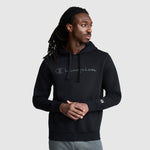 A man with long dreadlocks wears a black Champion Powerblend Hoodie, Tonal Script Logo, paired with gray sweatpants. He stands against a plain light gray background, looking to his right with his hands clasped.