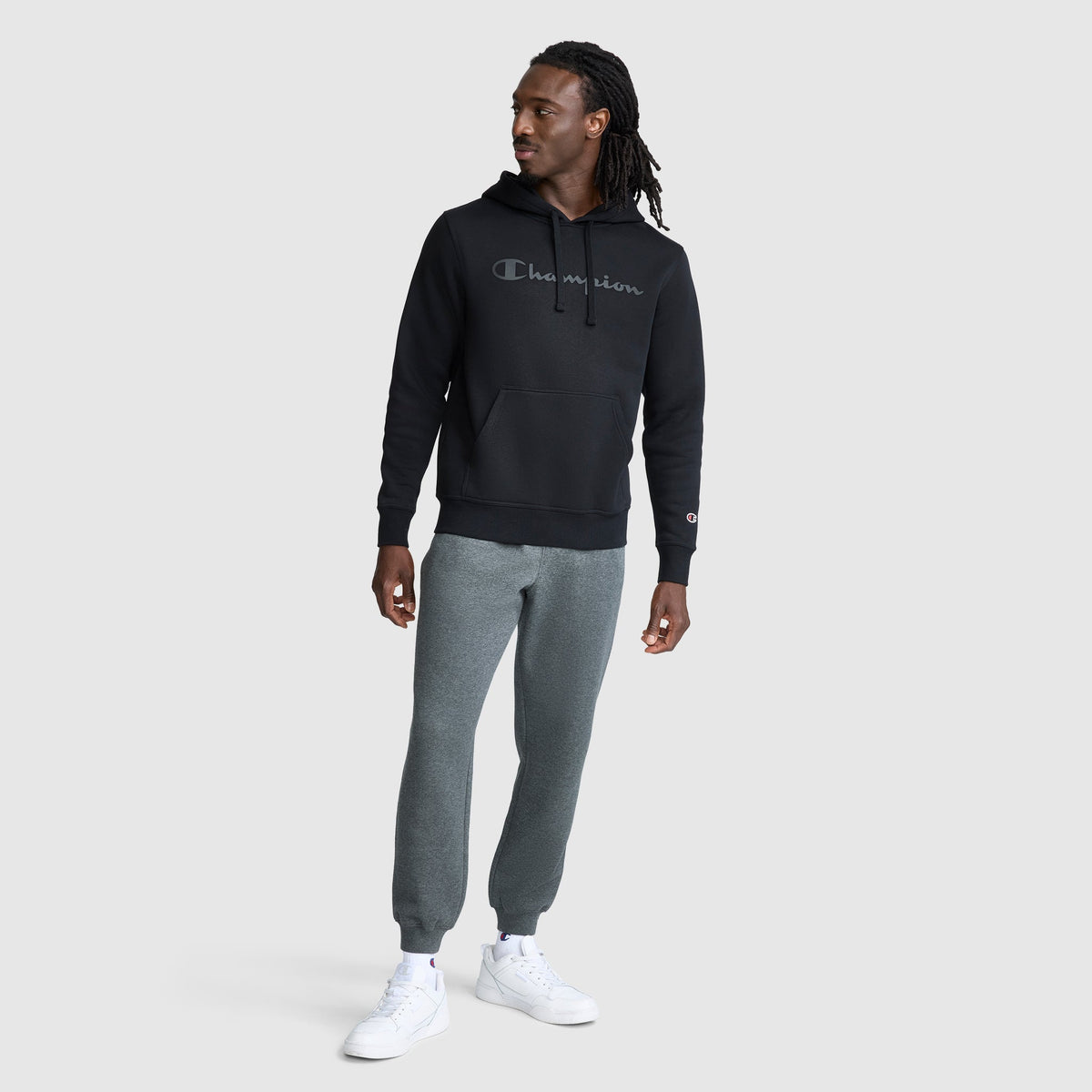 A person wearing a black Champion Powerblend Hoodie, Tonal Script Logo with a tonal classic script logo across the chest, paired with gray sweatpants and white sneakers.