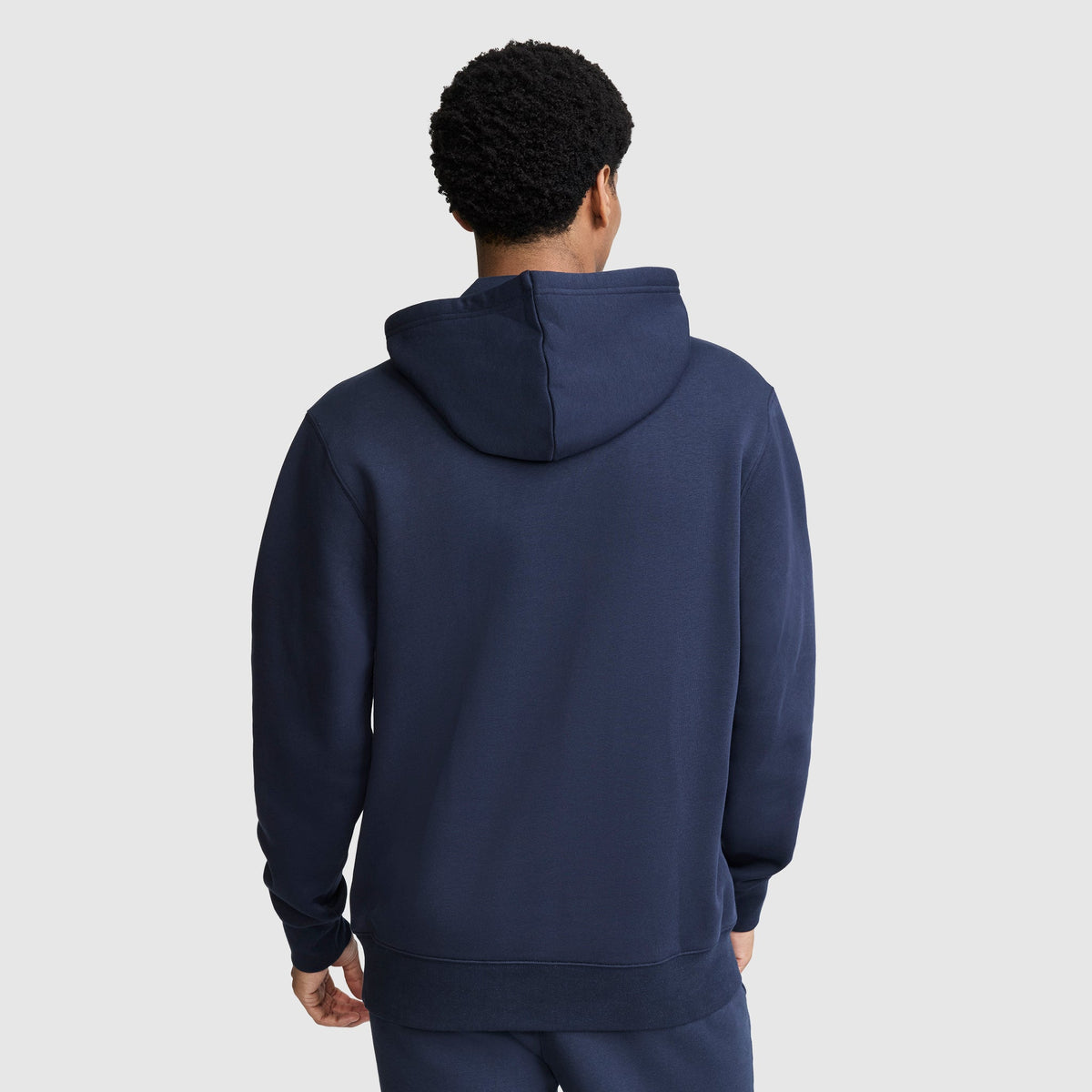 A person stands facing away, wearing a Champion Powerblend Hoodie with a tonal script logo in navy blue and matching navy pants. The hood is down, and the plain light gray background highlights the hoodie’s classic style.