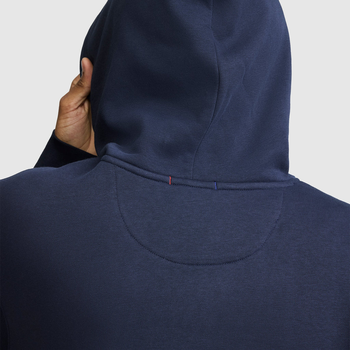 A person wearing a navy blue Champion Powerblend Hoodie with a tonal script logo is seen from behind, one hand touching the side of their hood. The fabric features visible stitching and two small colored threads near the neckline, characteristic of the brand’s iconic detailing.