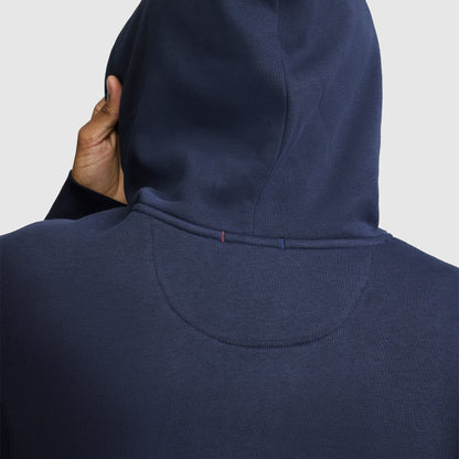 A person wearing a navy blue Champion Powerblend Hoodie with a tonal script logo is seen from behind, one hand touching the side of their hood. The fabric features visible stitching and two small colored threads near the neckline, characteristic of the brand’s iconic detailing.