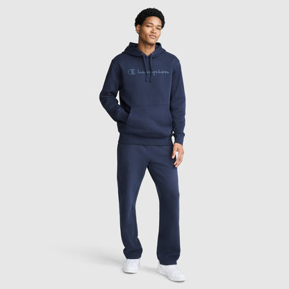 A person stands wearing a Champion Powerblend Hoodie with a tonal script logo in navy blue, paired with matching Powerblend Fleece sweatpants and white sneakers. They pose with one hand in the hoodie pocket and the other relaxed at their side against a plain light gray background.