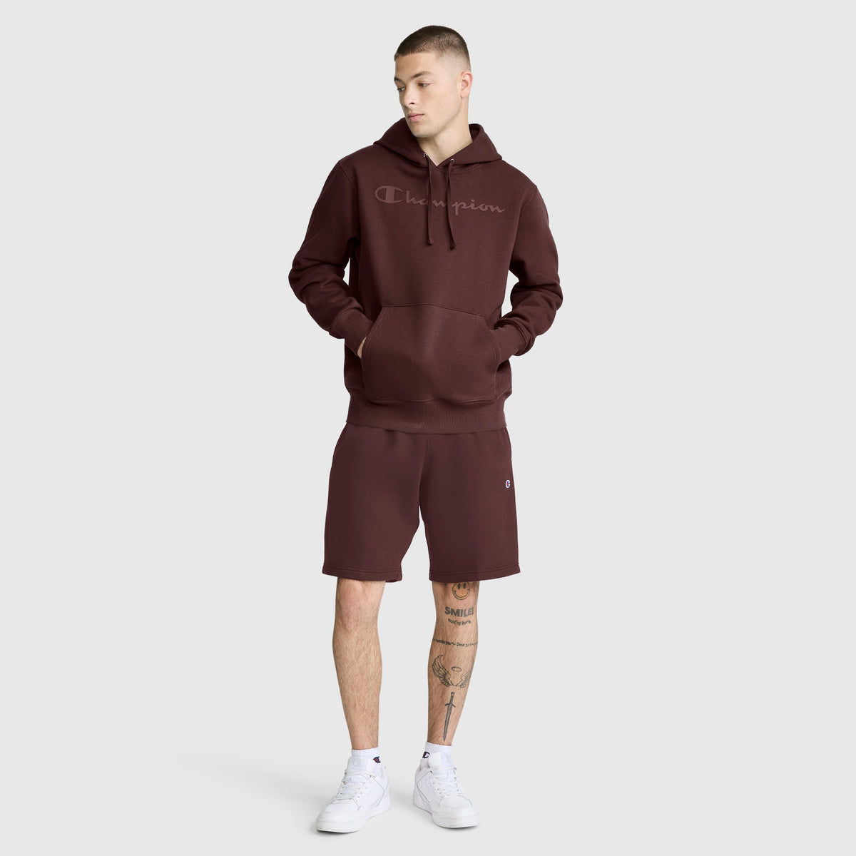 A person wearing a maroon Champion Powerblend Hoodie with Tonal Script Logo and matching shorts stands with hands in the hoodie pocket, looking to the side. The men’s hoodie, featuring Champion’s signature tonal script logo, is paired with white sneakers and leg tattoos for a stylish, laid-back look.