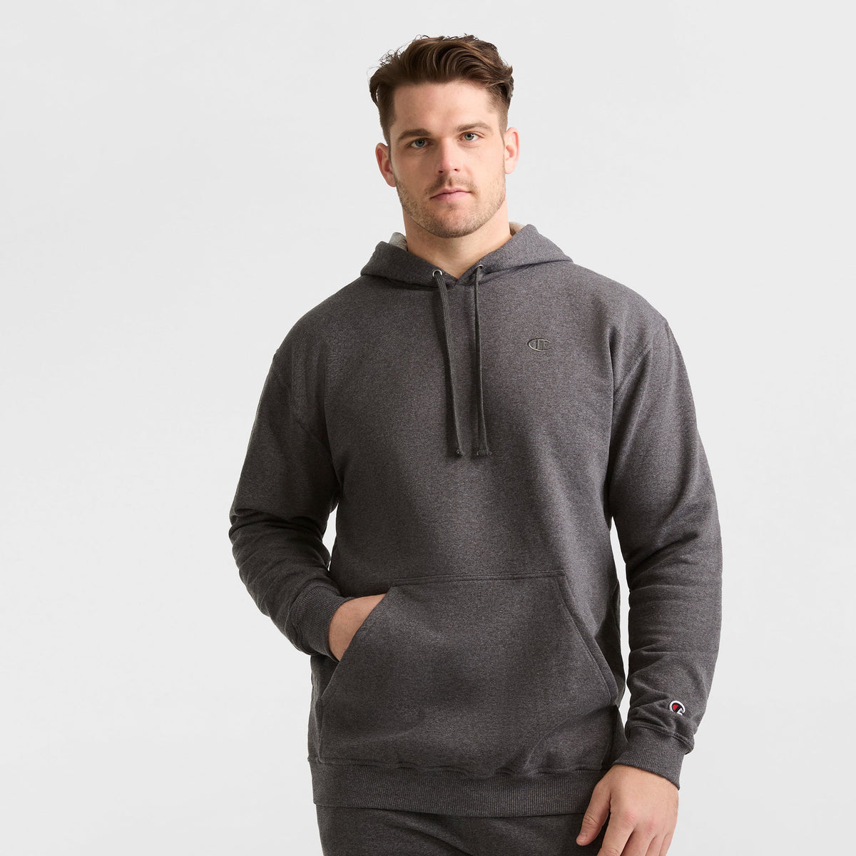 A man wearing a Champion Powerblend Hoodie, C Logo - Big & Tall in dark gray with a front pocket and drawstrings stands facing the camera against a plain light background.
