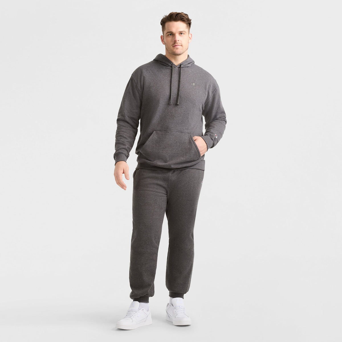 A man stands against a plain white background wearing the Champion Powerblend Hoodie, C Logo - Big & Tall in dark gray with matching sweatpants and white sneakers. One hand is in his pocket while the other rests by his side.