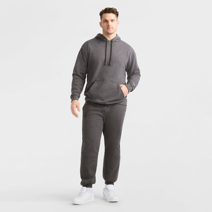 A man stands against a plain white background wearing the Champion Powerblend Hoodie, C Logo - Big & Tall in dark gray with matching sweatpants and white sneakers. One hand is in his pocket while the other rests by his side.