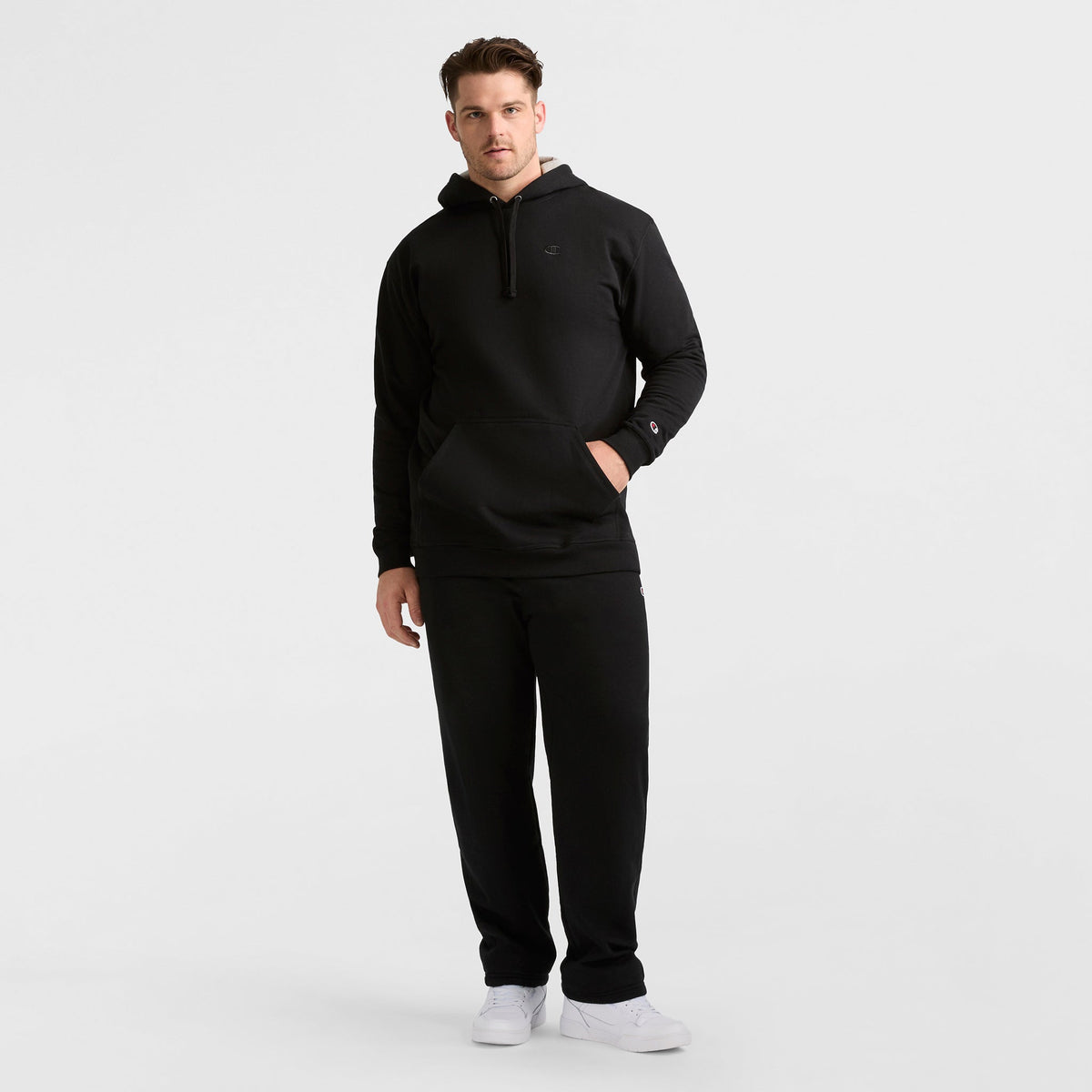 A man stands against a plain white background wearing a Champion Powerblend Hoodie, C Logo - Big & Tall in black with matching black sweatpants and white sneakers. One hand is in his hoodie pocket and he looks straight ahead.