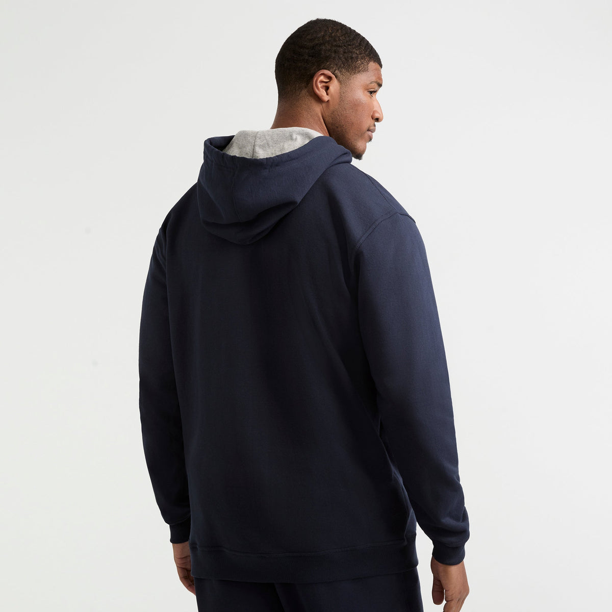 A man faces away from the camera, wearing a Champion Powerblend Hoodie, C Logo – Big & Tall in dark blue with the hood down. The background is plain white.