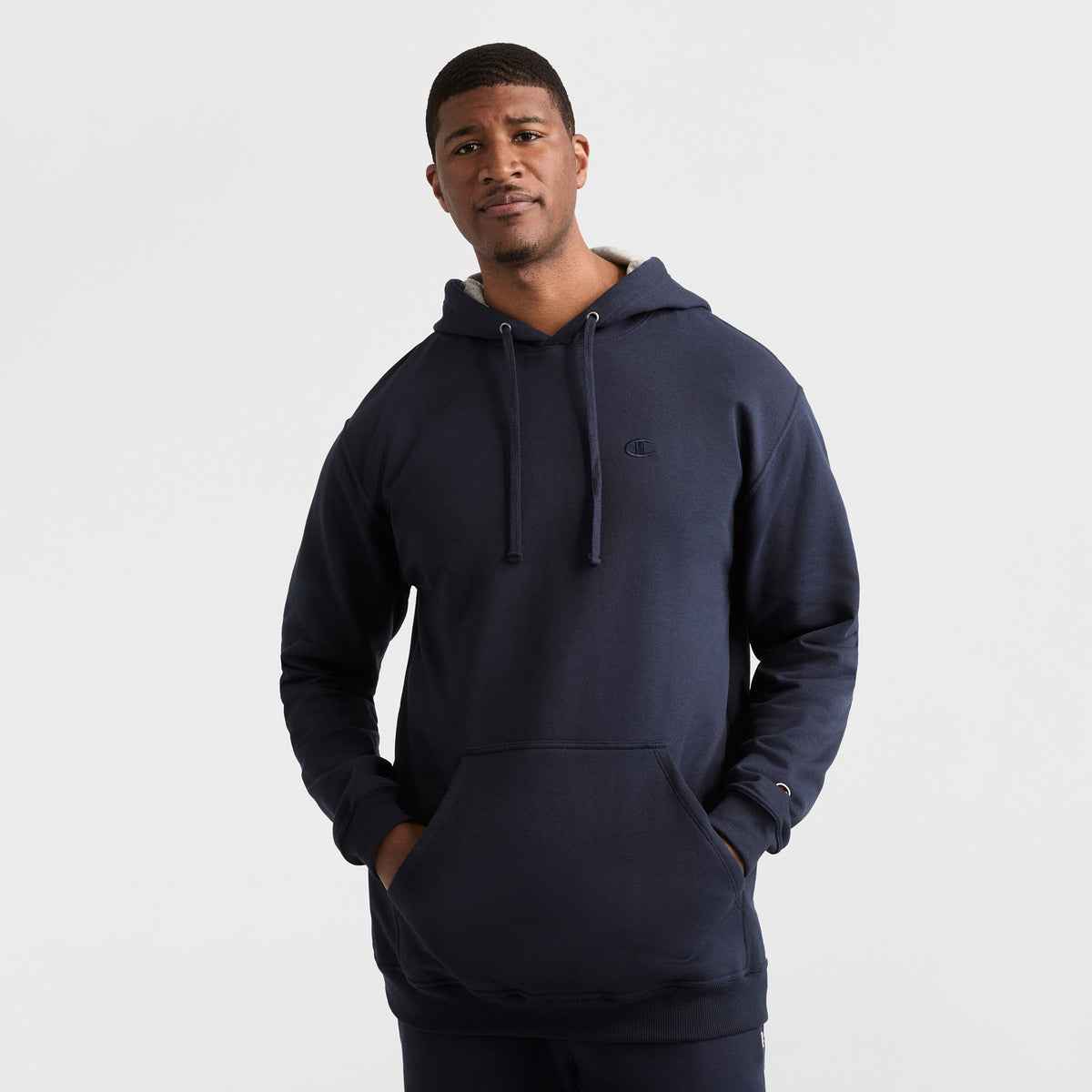 A man stands against a plain light background, wearing the Champion Powerblend Hoodie, C Logo - Big & Tall in navy blue with a front pocket and drawstrings. His hands are in the pocket, and he has a neutral expression.