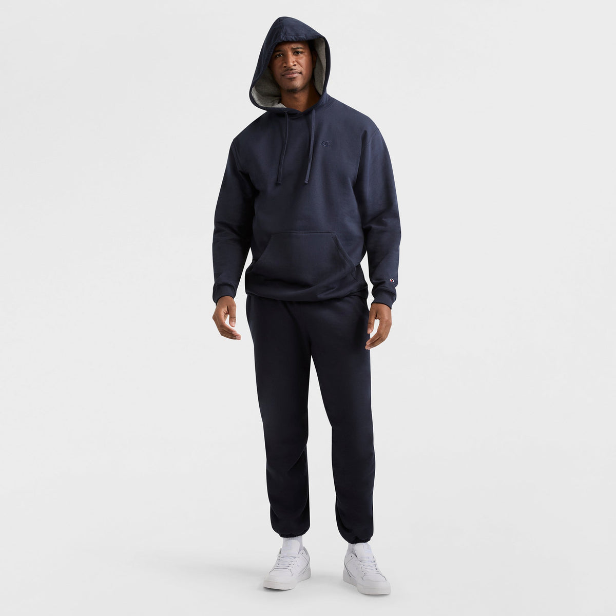 A man wears a Champion Powerblend Hoodie, C Logo - Big & Tall, in dark navy with matching sweatpants and white sneakers, hood up, standing facing forward against a plain white background.