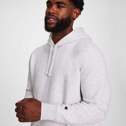A man wears a light gray Champion Powerblend Hoodie, C Logo - Big & Tall, standing against a white background and smiling slightly. The men's fleece hooded sweatshirt displays the Champion logo on his chest and sleeve.