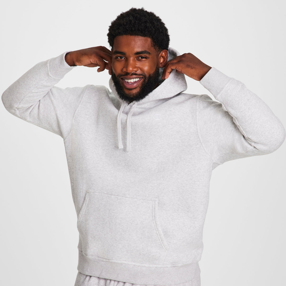 A bearded man smiles while wearing a light gray Champion Powerblend Hoodie, C Logo - Big & Tall, pulling on the hood with both hands against a plain white background.