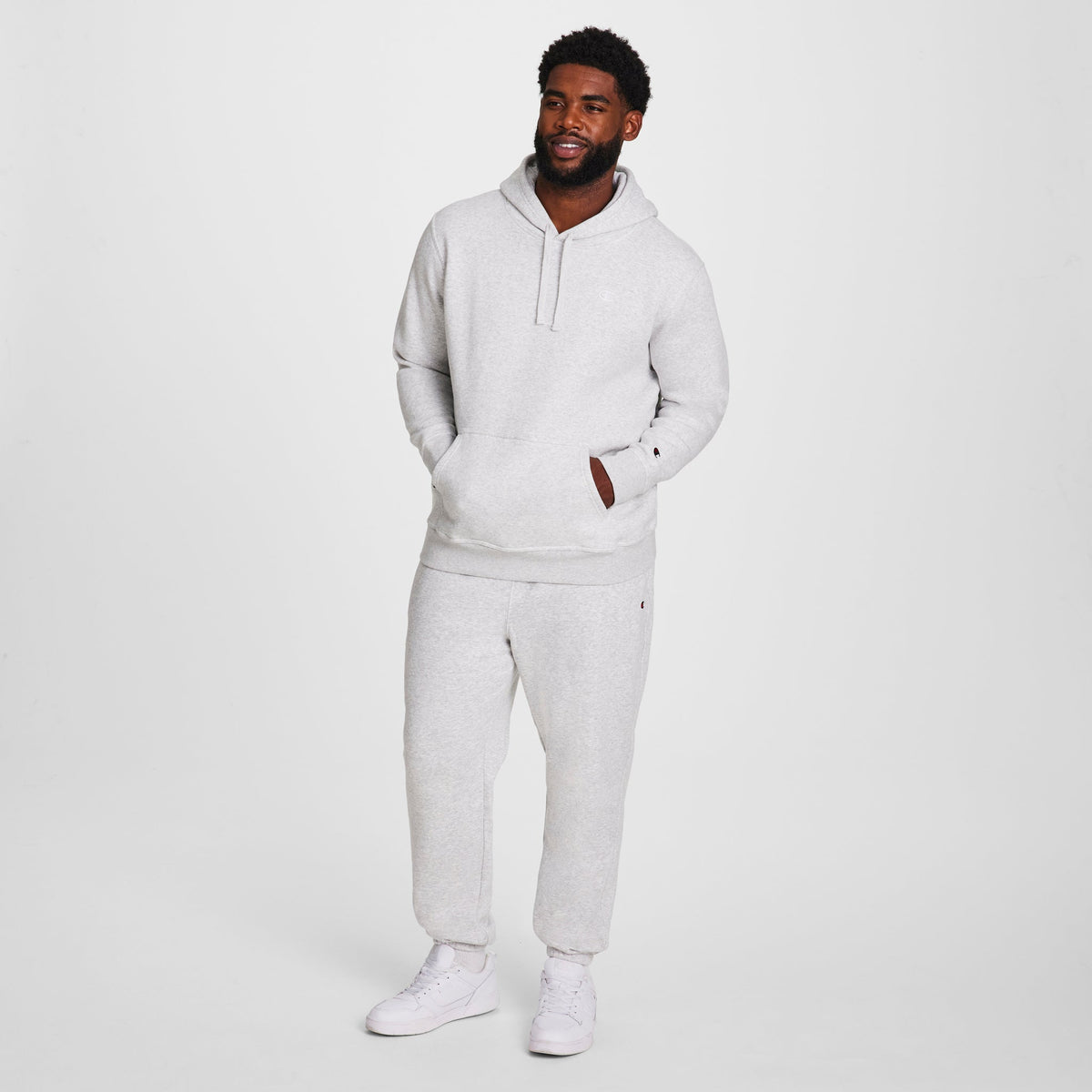 A man stands against a white background, wearing a Champion Powerblend Hoodie, C Logo - Big & Tall in light gray with matching sweatpants and white sneakers. His left hand is in his pocket and he smiles slightly.